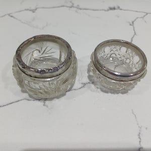 May include: Two clear glass jars with silver rims. The jars have a cut glass design and are of different sizes. The larger jar has a starburst pattern on the inside. The jars are on a white and grey marble surface.