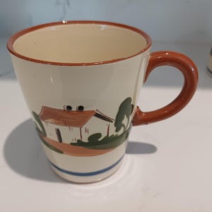 May include: Cream ceramic mug with a brown handle and rim. The mug has a hand-painted design of a house with a brown roof, green trees, and an orange base. A blue stripe accents the bottom.
