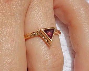 May include: Gold ring with a triangular red gemstone set in a gold setting. The ring is on a finger.