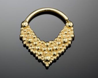 May include: A gold-colored septum ring with a decorative, beaded design. The ring has a hinged closure and a V-shaped beaded section. The beads are small and closely spaced, creating a textured surface. The jewelry is displayed on a reflective black surface.