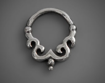May include: Silver septum ring with a decorative, ornate design. The ring has a circular shape with a detailed, swirling pattern and a small bead at the bottom.