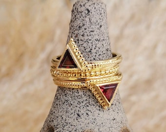 May include: Gold ring with three bands and two red triangular gemstones. The ring is on a gray textured display stand.