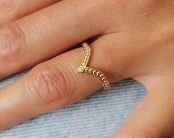 May include: A gold ring with a V-shaped design and a small diamond in the center. The ring is adorned with small beads along the edges.
