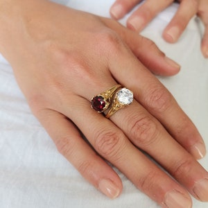 May include: A gold ring with a red and a clear gemstone. The ring is on a finger.