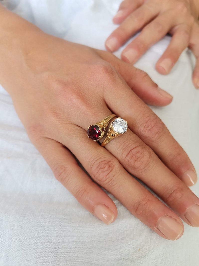 May include: A gold ring with a red and a clear gemstone. The ring is on a finger.