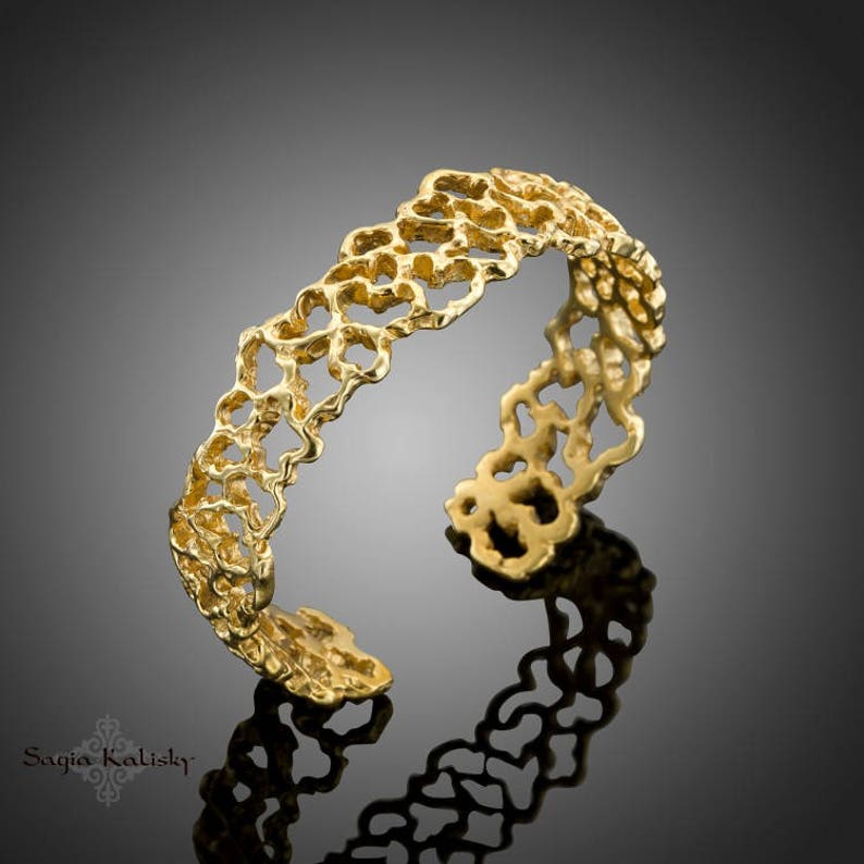 May include: A gold-toned ring with a delicate, intricate design. The ring is made of a thin, openwork band with a repeating pattern of small, rounded shapes. The ring is shown on a gray background.