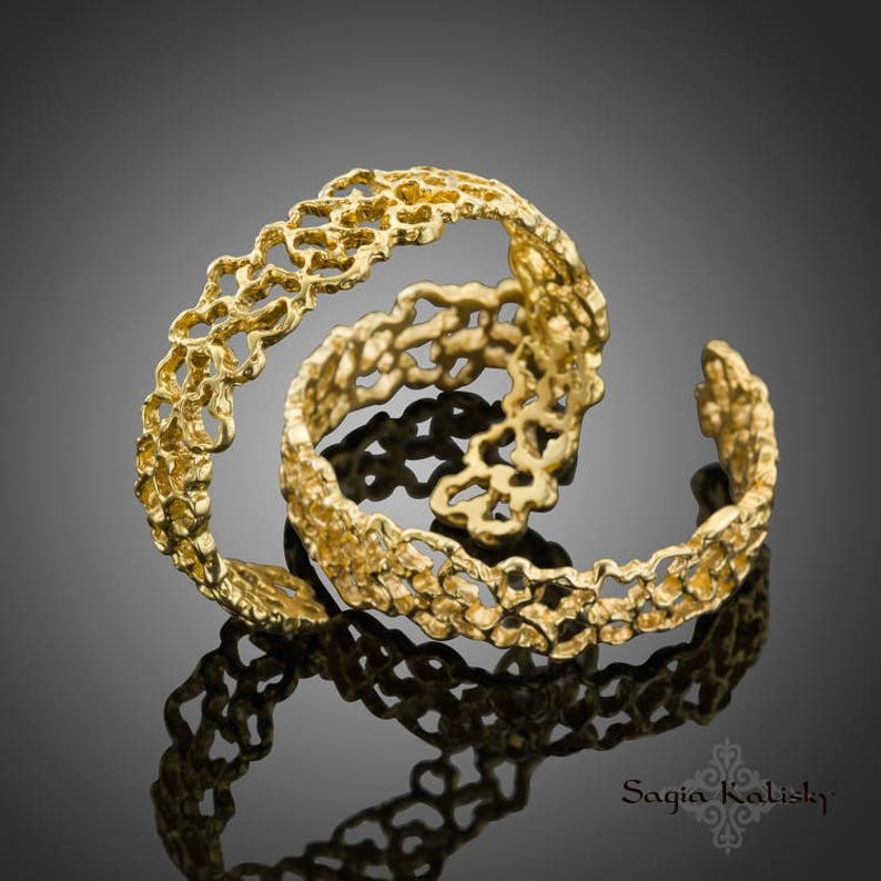 May include: Two gold rings with a delicate, intricate design resembling a floral pattern. The rings are stacked on top of each other, creating a layered effect.