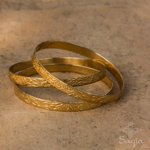 May include: Three gold-colored metal bangles with an intricate floral design. The bangles are stacked on top of each other.