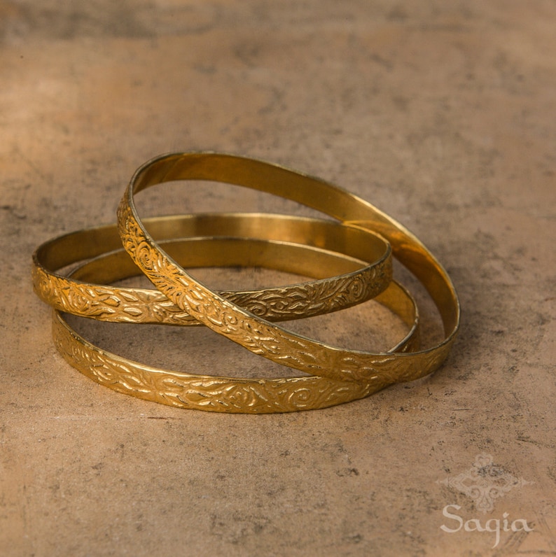 May include: Three gold-colored metal bangles with an intricate floral design. The bangles are stacked on top of each other.