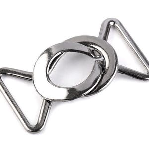 May include: Silver metal buckle with a D-ring and two triangular loops. The buckle is designed for attaching straps or belts.
