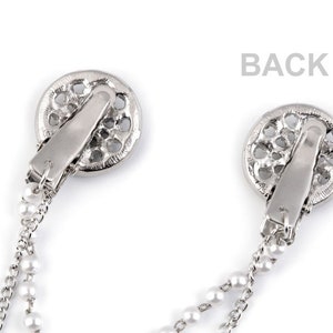 May include: A pair of silver tone jewelry clips with pearl chains. Each clip features a circular design with an openwork pattern.