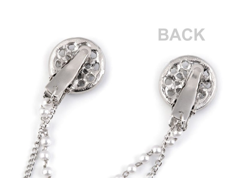 May include: A pair of silver tone jewelry clips with pearl chains. Each clip features a circular design with an openwork pattern.