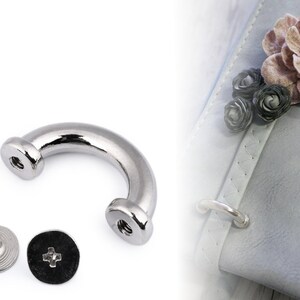 May include: Silver metal D-ring hardware with two screws. The D-ring is a U-shaped piece of metal with a rounded top and two screw holes on the sides. The screws are silver metal with a Phillips head.