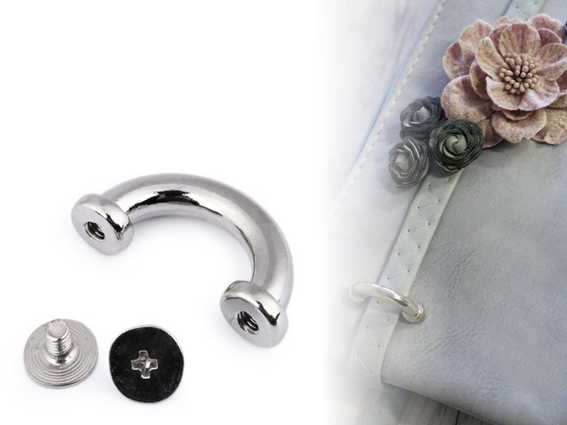 May include: Silver metal D-ring hardware with two screws. The D-ring is a U-shaped piece of metal with a rounded top and two screw holes on the sides. The screws are silver metal with a Phillips head.