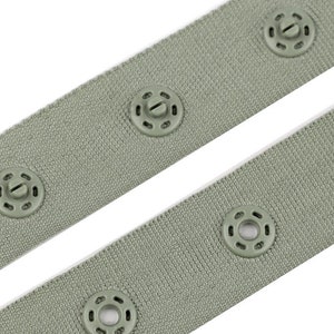 Puede incluir: Primer plano de dos correas de tela verde salvia con cierres de presi&oacute;n a juego y espaciados uniformemente. Los broches y la tela comparten un color similar, creando un aspecto cohesivo. La tela tiene un aspecto texturizado.