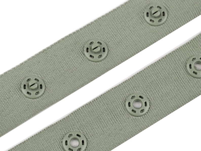 Puede incluir: Primer plano de dos correas de tela verde salvia con cierres de presi&oacute;n a juego y espaciados uniformemente. Los broches y la tela comparten un color similar, creando un aspecto cohesivo. La tela tiene un aspecto texturizado.
