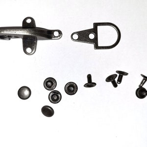 May include: A set of silver-toned metal hardware for leather crafting. The set includes a hook and loop closure, a D-ring, and several rivets.
