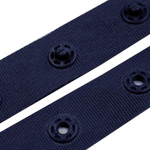 Puede incluir: Primer plano de dos correas de tela azul marino con broches negros. Las correas est&aacute;n en &aacute;ngulo diagonal, mostrando la textura de la tela y los detalles de los broches. Los broches est&aacute;n espaciados uniformemente a lo largo de las correas.