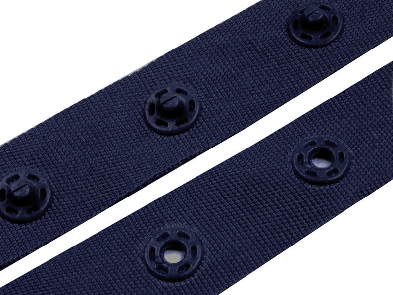 Puede incluir: Primer plano de dos correas de tela azul marino con broches negros. Las correas est&aacute;n en &aacute;ngulo diagonal, mostrando la textura de la tela y los detalles de los broches. Los broches est&aacute;n espaciados uniformemente a lo largo de las correas.