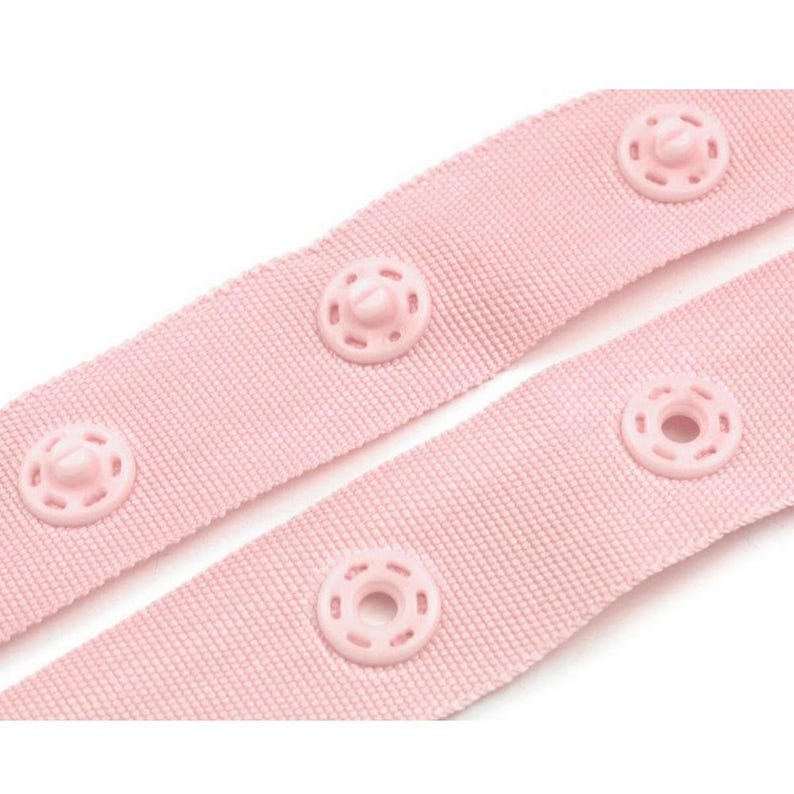 Puede incluir: Primer plano de dos tiras de tela rosa claro con broches rosas a juego y espaciados uniformemente. La tela tiene un tejido texturizado, y los broches son circulares con un bot&oacute;n central.