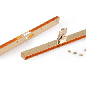 Op de afbeelding: Twee gouden metalen frames met een gouden metalen sluiting en kleine gouden schroeven. De frames zijn rechthoekig en hebben een gleuf in het midden. De sluiting heeft een klein wiel aan de zijkant.