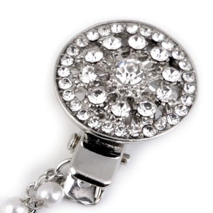 May include: A silver-toned brooch with a round, crystal-encrusted design. The brooch has a clip on the back and a chain with pearls attached.