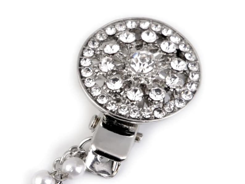 May include: A silver-toned brooch with a round, crystal-encrusted design. The brooch has a clip on the back and a chain with pearls attached.