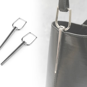 May include: Two silver metal bag hooks with a rectangular loop at the top and a pointed end. The hooks are shown against a white background.