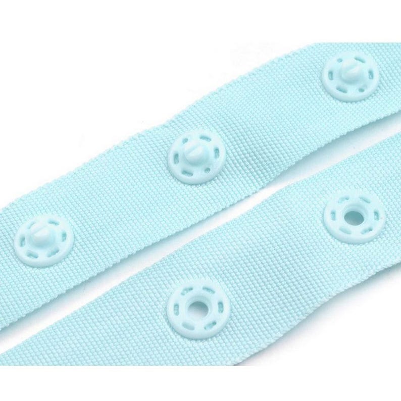 Puede incluir: Primer plano de dos correas de tela azul claro con broches blancos. Las correas est&aacute;n hechas de un material tejido y tienen cuatro broches blancos espaciados uniformemente a lo largo de su longitud. Los broches son circulares con un bot&oacute;n central y agujeros circundantes.