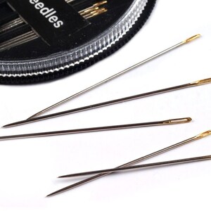 May include: A close-up of four silver sewing needles with gold eye tips. The needles are arranged in a fan shape on a white surface.