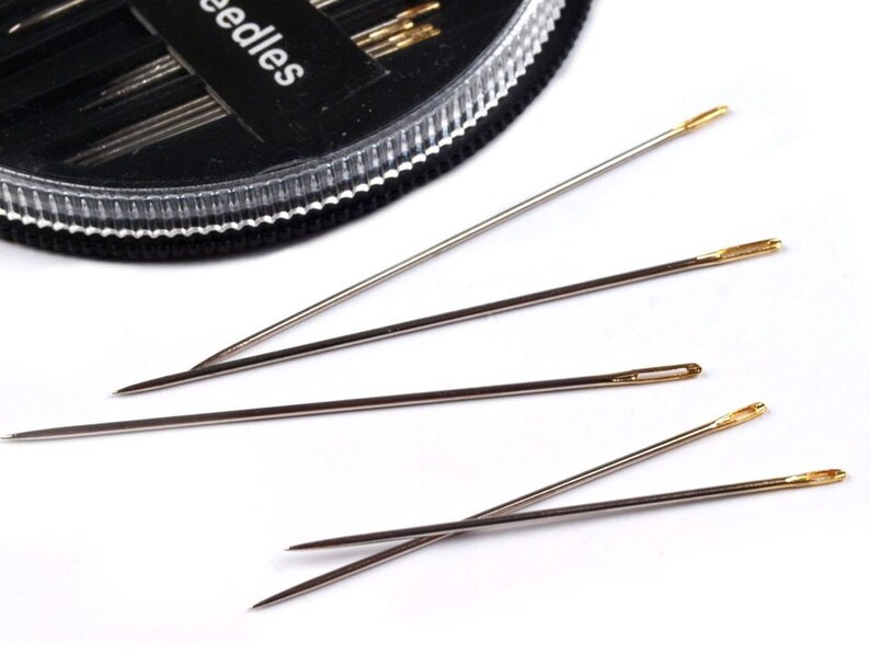 May include: A close-up of four silver sewing needles with gold eye tips. The needles are arranged in a fan shape on a white surface.