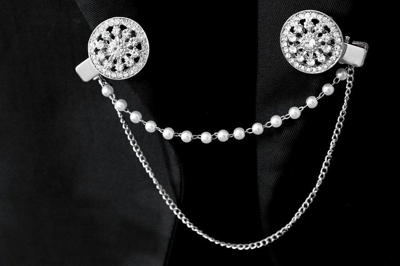 May include: A silver chain with a pearl strand and two round, silver, rhinestone-encrusted brooches.