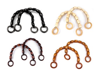 May include: Four pairs of wooden bag handles in various colors: black, natural, dark brown, and medium brown. Each handle is made of oval beads strung together, with a circular ring at each end for attachment. The handles are arranged on a white background.