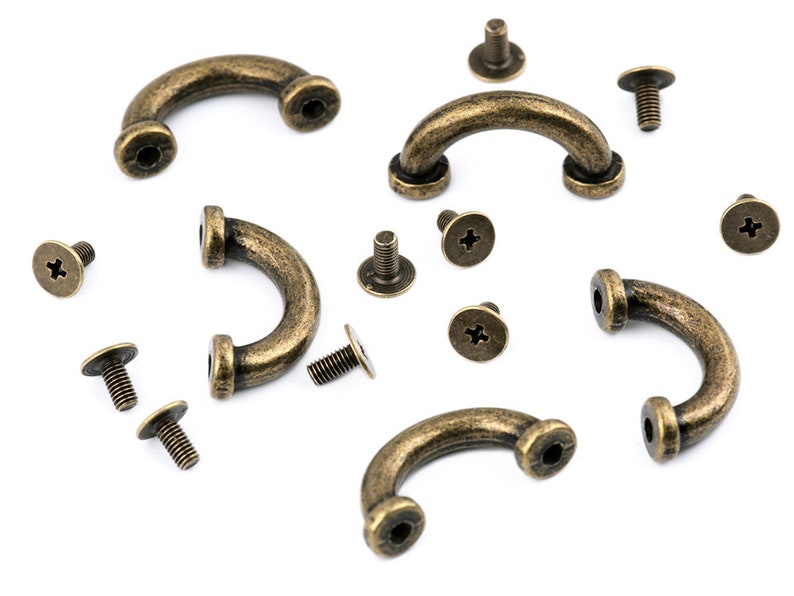 May include: A set of antique brass-colored metal handles with screws. The handles are shaped like a half-circle and have a smooth, slightly textured surface. The screws are small and have a cross-shaped head.