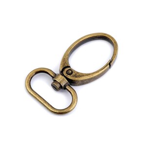 May include: A gold-toned metal swivel clasp with a D-ring. The clasp is open and ready to be attached to a strap or chain.