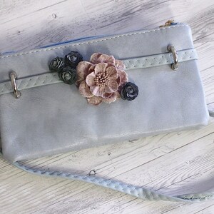 May include: A light blue leather clutch with a braided strap and a floral embellishment. The flowers are pink, black, and white.