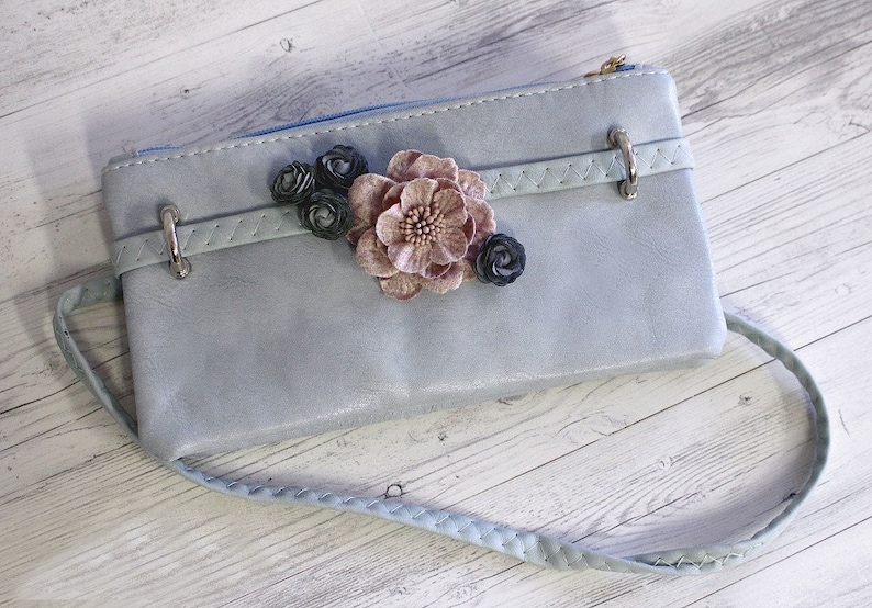 May include: A light blue leather clutch with a braided strap and a floral embellishment. The flowers are pink, black, and white.