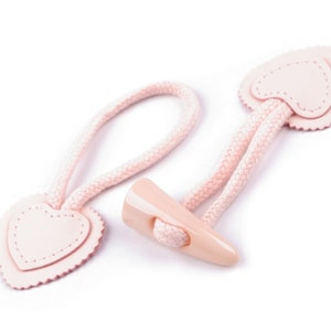 Pode incluir: Um par de botões de coração rosa com um cordão rosa e um botão de plástico. Os botões são costurados com um ponto decorativo.