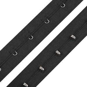 Peut inclure: Deux bandes de ruban noir avec crochets et œillets. Chaque bande comporte plusieurs crochets et œillets métalliques de couleur argentée. Les bandes sont en tissu tissé et sont inclinées en diagonale sur un fond blanc.