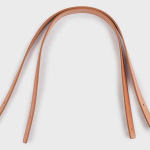 May include: Two light brown leather bag handles with a curved design. Each handle has two holes at the ends for attachment. The handles are made of a smooth, natural-looking leather.