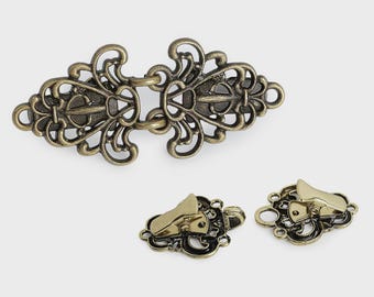 May include: A bronze-toned metal cloak clasp featuring an intricate filigree design with two symmetrical halves that hook together, accompanied by two smaller matching metal clips with a polished finish and ornate cut-out detailing.