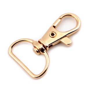 May include: A gold-toned metal swivel clasp with a D-ring. The clasp is open and ready to be attached to a strap or chain.