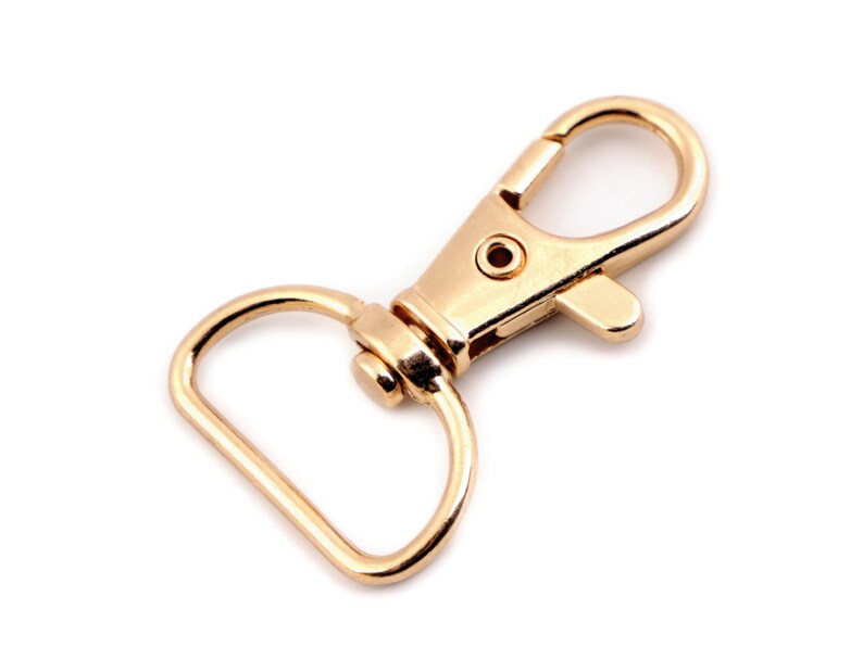 May include: A gold-toned metal swivel clasp with a D-ring. The clasp is open and ready to be attached to a strap or chain.