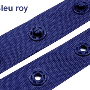 Puede incluir: Primer plano de dos correas de tela azul real con broches de presi&oacute;n azul oscuro. Las correas est&aacute;n colocadas en diagonal sobre un fondo blanco. El texto "Bleu roy" est&aacute; en la esquina superior izquierda.
