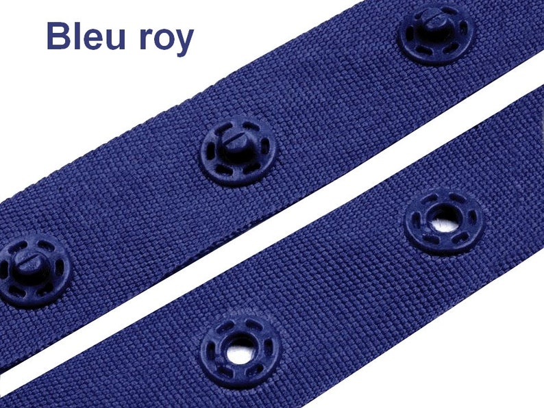 Puede incluir: Primer plano de dos correas de tela azul real con broches de presi&oacute;n azul oscuro. Las correas est&aacute;n colocadas en diagonal sobre un fondo blanco. El texto "Bleu roy" est&aacute; en la esquina superior izquierda.