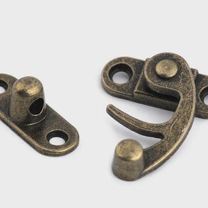 May include: Two antique bronze-colored metal clasps. One is a rectangular plate with a rounded post and two holes. The other is a hinged clasp with a hook and two holes. Hardware for boxes, chests, or crafts.