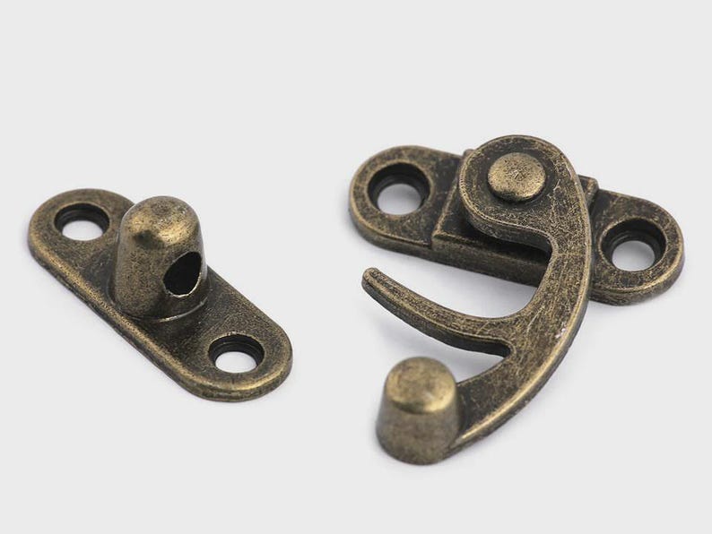May include: Two antique bronze-colored metal clasps. One is a rectangular plate with a rounded post and two holes. The other is a hinged clasp with a hook and two holes. Hardware for boxes, chests, or crafts.