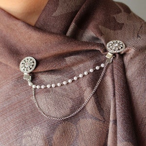 May include: A pair of silver scarf clips with rhinestone accents and a pearl chain connecting them. The clips are attached to a brown patterned scarf.