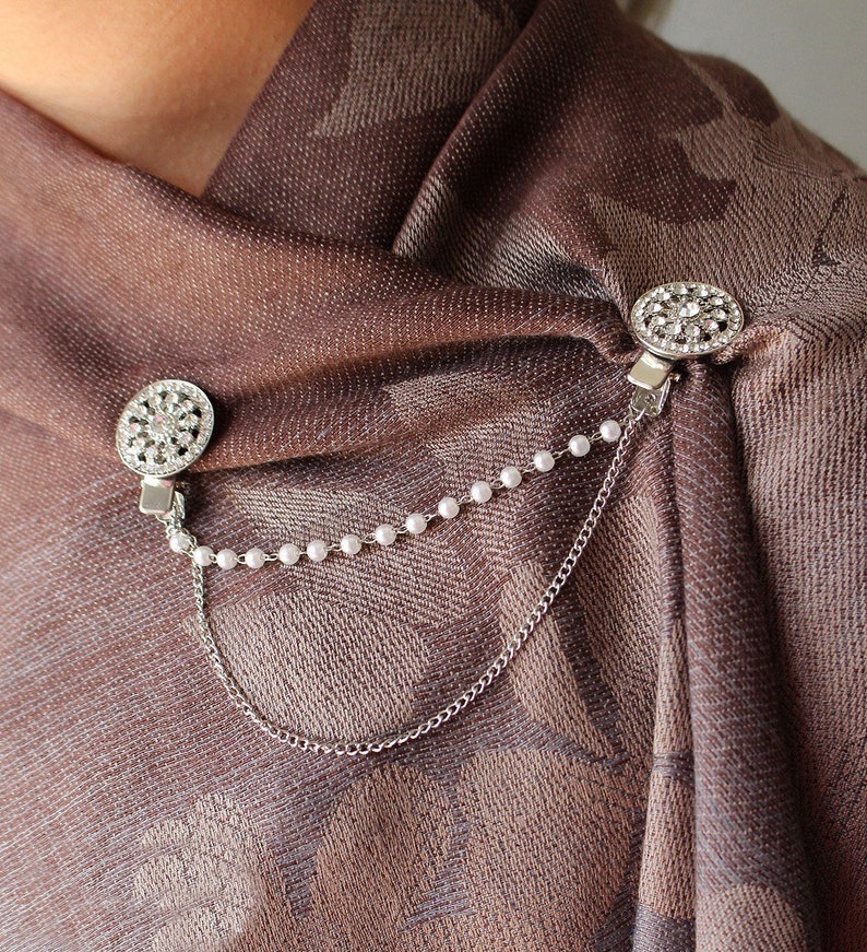 May include: A pair of silver scarf clips with rhinestone accents and a pearl chain connecting them. The clips are attached to a brown patterned scarf.