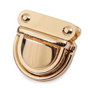 Pode incluir: Fecho de metal dourado com um design arredondado e articulado. O fecho tem duas pequenas maçanetas redondas na parte superior.